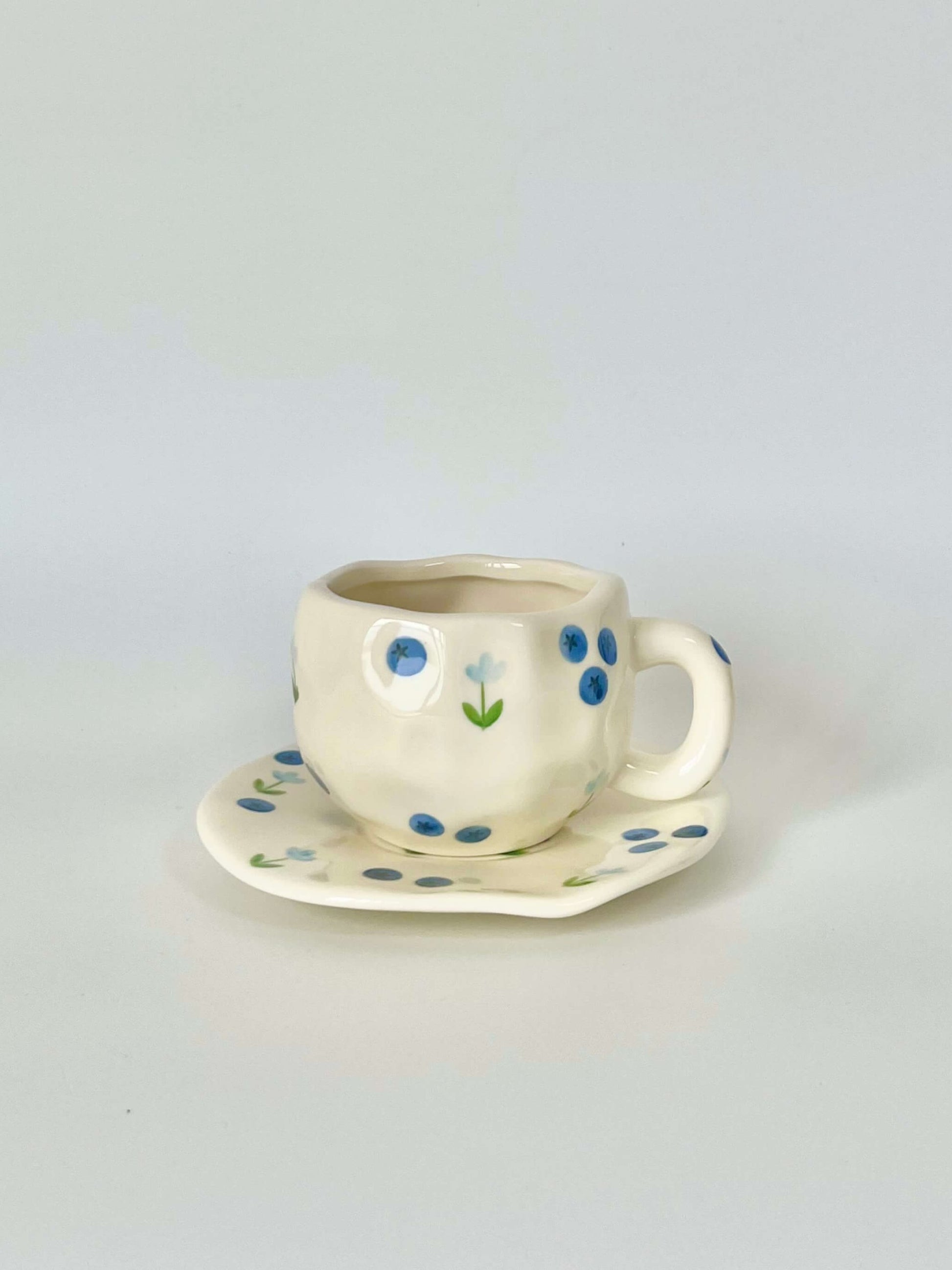 Cute blue cherry tea set with mug and saucer together. Adorable beige background with hand painted blueberry accents drawing on. Coquette girl aesthetic. Perfect gift for mums, daughters and girlfriends. Cafe vibes at home.