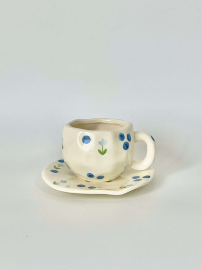 Cute blue cherry tea set with mug and saucer together. Adorable beige background with hand painted blueberry accents drawing on. Coquette girl aesthetic. Perfect gift for mums, daughters and girlfriends. Cafe vibes at home.