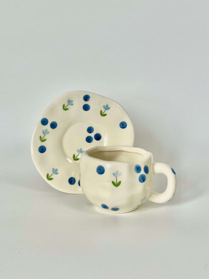 Cute blue cherry tea set with mug and saucer together. Adorable beige background with hand painted blueberry accents drawing on. Coquette girl aesthetic. Perfect gift for mums, daughters and girlfriends. Cafe vibes at home.