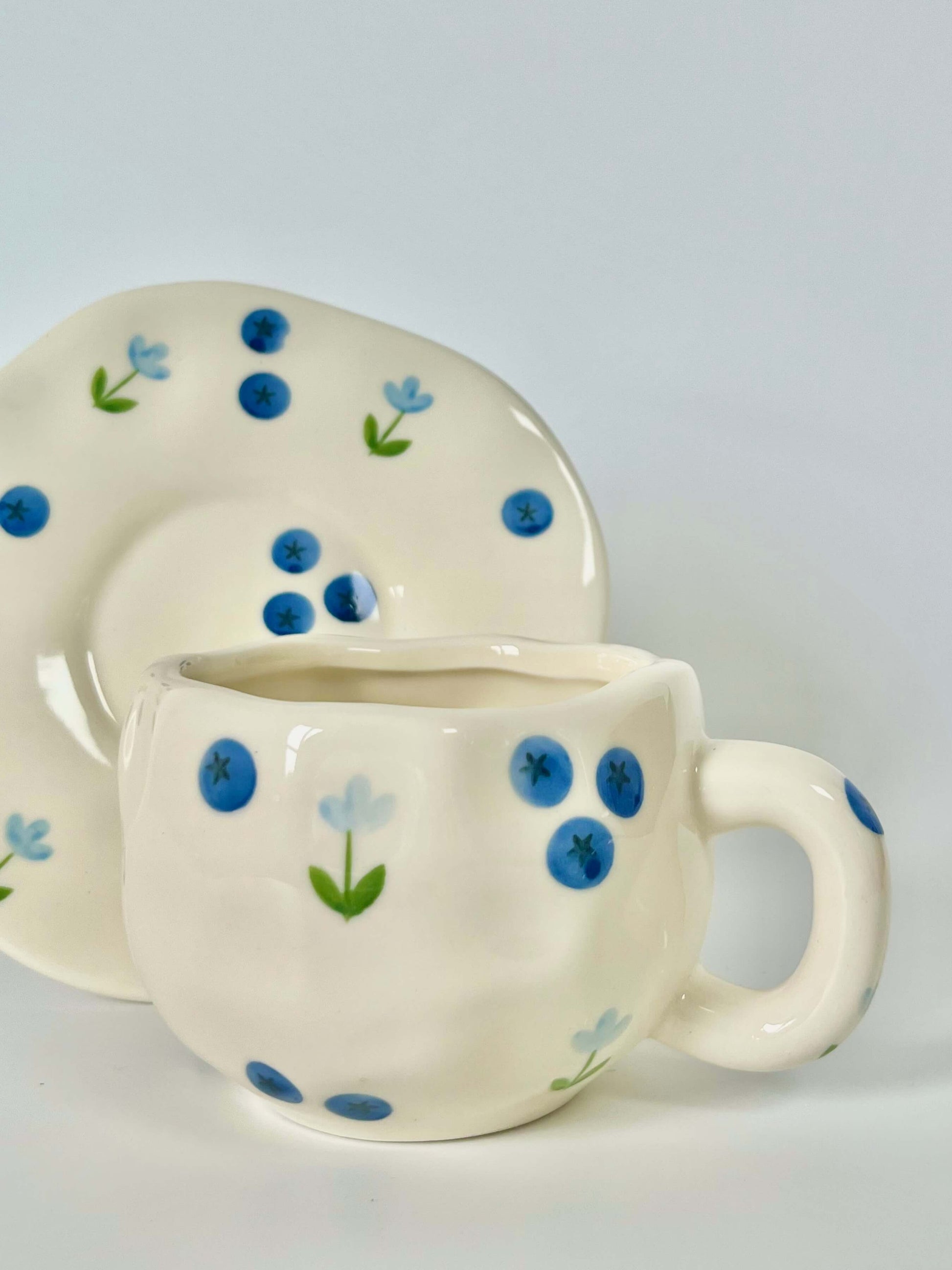 Cute blue cherry tea set with mug and saucer together. Adorable beige background with hand painted blueberry accents drawing on. Coquette girl aesthetic. Perfect gift for mums, daughters and girlfriends. Cafe vibes at home.