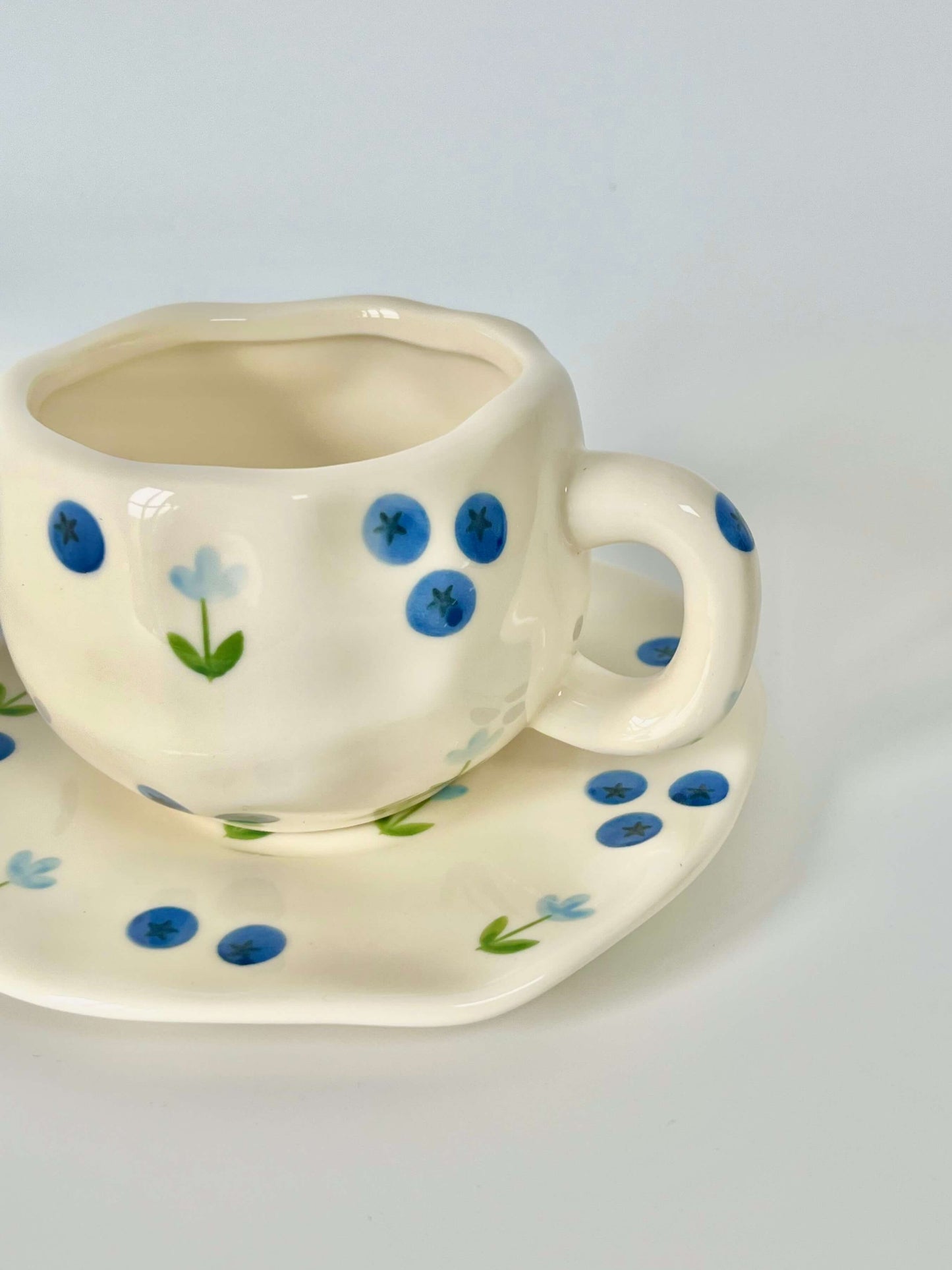 Cute blue cherry tea set with mug and saucer together. Adorable beige background with hand painted blueberry accents drawing on. Coquette girl aesthetic. Perfect gift for mums, daughters and girlfriends. Cafe vibes at home.