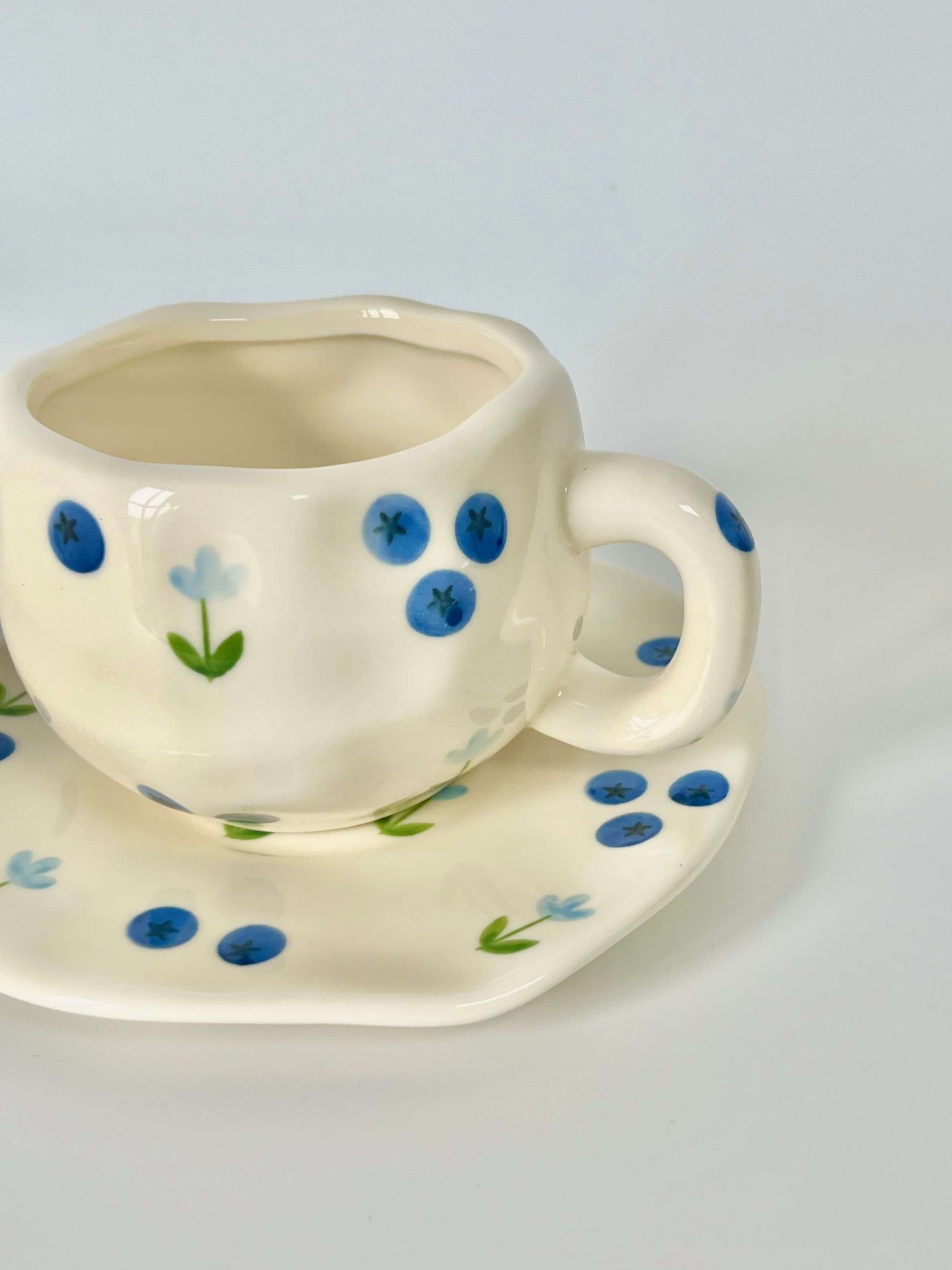 Cute blue cherry tea set with mug and saucer together. Adorable beige background with hand painted blueberry accents drawing on. Coquette girl aesthetic. Perfect gift for mums, daughters and girlfriends. Cafe vibes at home.