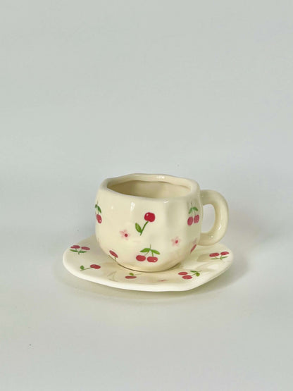 Cute blue cherry tea set with mug and saucer together. Adorable beige background with hand painted red cherry accents drawing on. Coquette girl aesthetic. Perfect gift for mums, daughters and girlfriends. Cafe vibes at home.