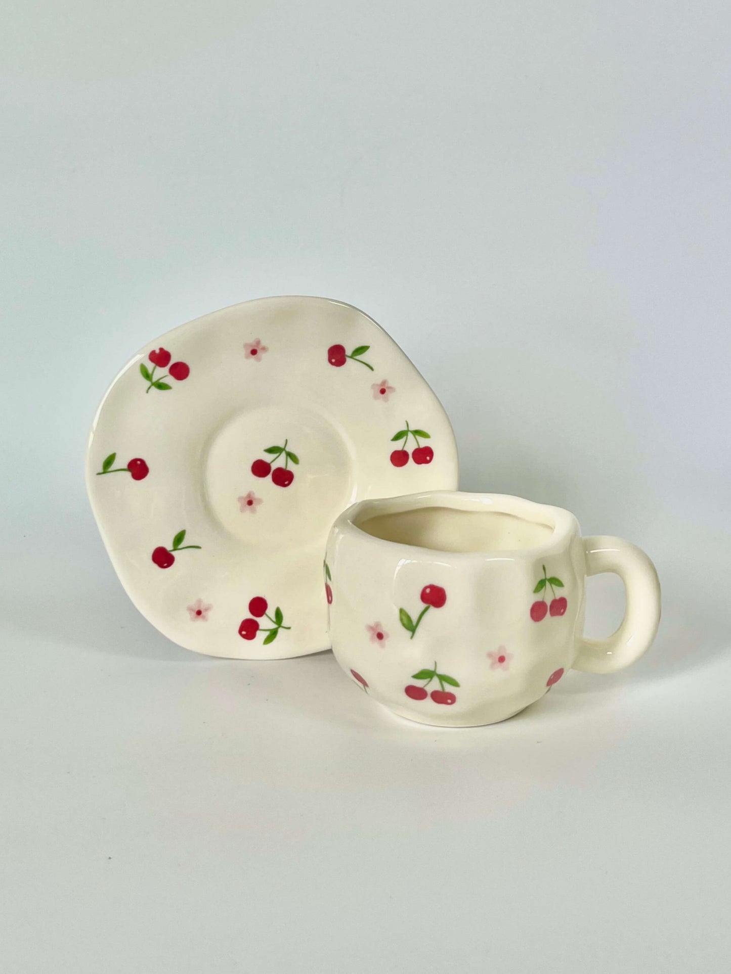 Cute blue cherry tea set with mug and saucer together. Adorable beige background with hand painted red cherry accents drawing on. Coquette girl aesthetic. Perfect gift for mums, daughters and girlfriends. Cafe vibes at home.