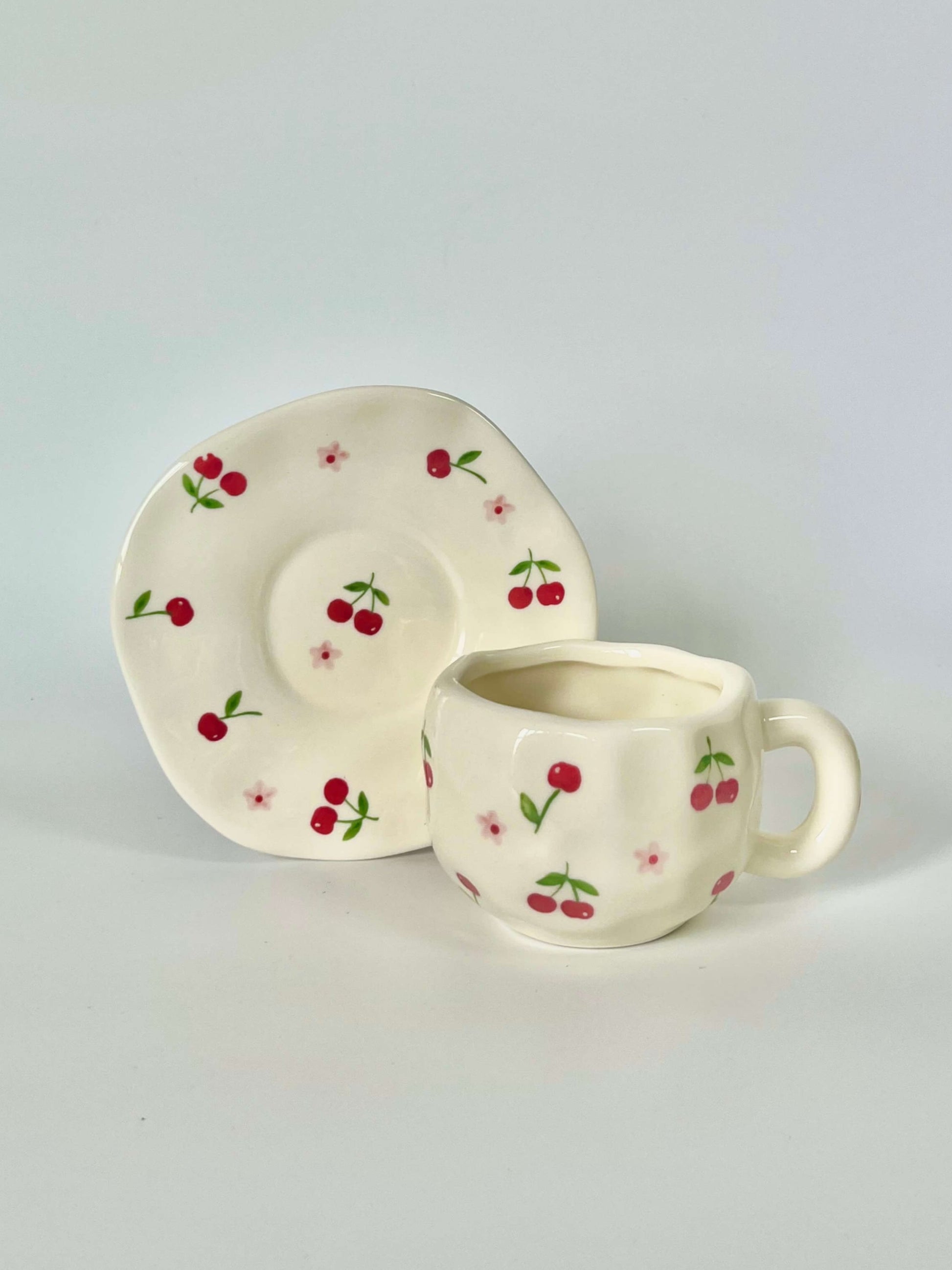 Cute blue cherry tea set with mug and saucer together. Adorable beige background with hand painted red cherry accents drawing on. Coquette girl aesthetic. Perfect gift for mums, daughters and girlfriends. Cafe vibes at home.