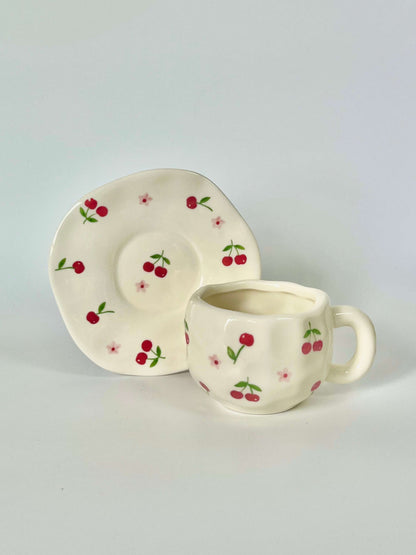 Cute blue cherry tea set with mug and saucer together. Adorable beige background with hand painted red cherry accents drawing on. Coquette girl aesthetic. Perfect gift for mums, daughters and girlfriends. Cafe vibes at home.